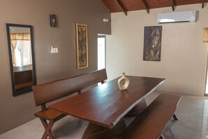 A dining area with a wooden bench, a table, and a vase. There are framed artworks on the walls and an air conditioning unit above.