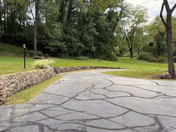 Un camino de entrada pavimentado con borde de piedra, rodeado de césped y árboles.