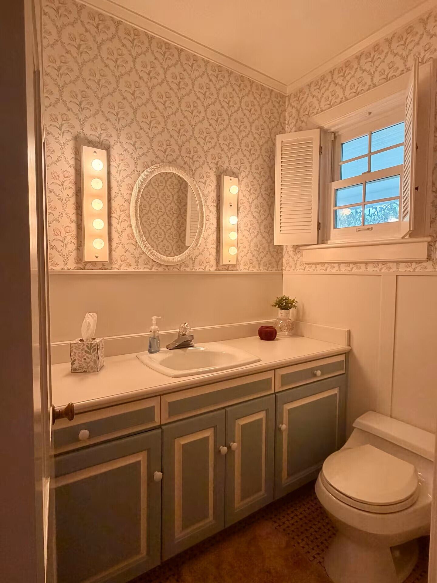 A bathroom with a vanity, a mirror with lights, a toilet, and a window with shutters.