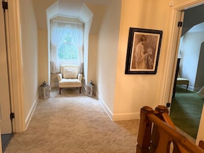 Un pasillo con piso alfombrado, una silla blanca, un cuadro en la pared y una puerta que da a otra habitación.