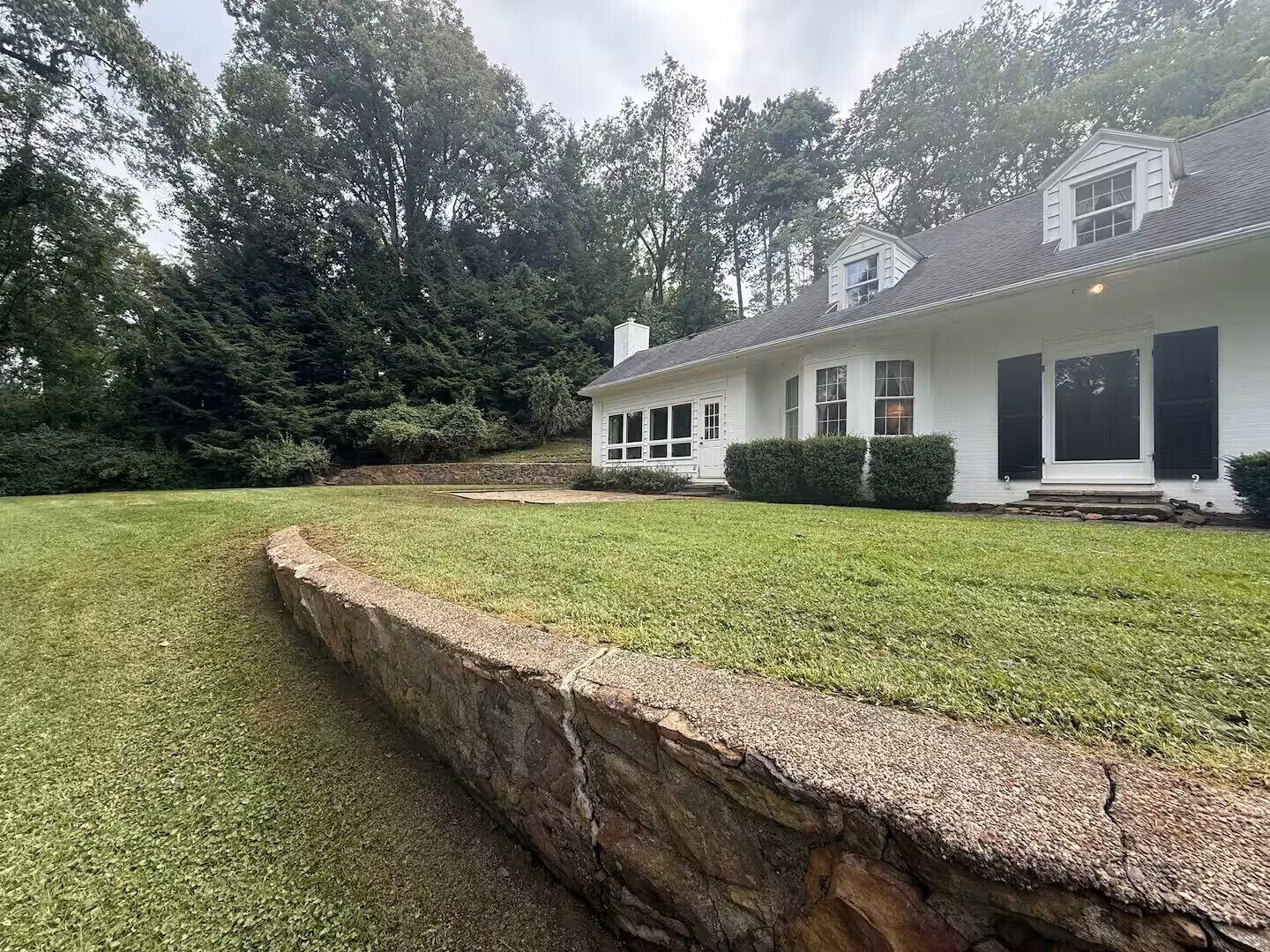 A single-story house with a stone retaining wall, a lawn, and a dense forest in the background.