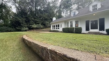 A single-story house with a stone retaining wall, a lawn, and a dense forest in the background.