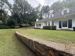 Una casa de un piso con un muro de contención de piedra, césped y un bosque frondoso al fondo.