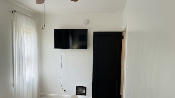 A bedroom with a bed, a television mounted on the wall, a black door, and a ceiling fan.