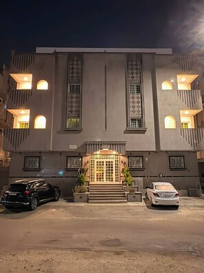 Ein Gebäude mit einer symmetrischen Fassade, einem zentralen Eingang mit Stufen und zwei geparkten Autos davor.