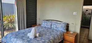 Ein Bett mit floralem Überwurf, zwei Kissen, ein zusammengerolltes Handtuch, ein Nachttisch, eine Lampe und Blick auf einen Garten durch ein Fenster.