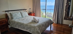 Ein Bett mit floralem Überwurf, ein Nachttisch, Holzboden, Meerblick durch ein Fenster und ein Spiegel an der Wand.