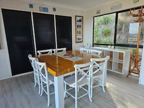 Un comedor con mesa de madera y sillas blancas, una puerta negra y un ventanal con vistas a la vegetación.