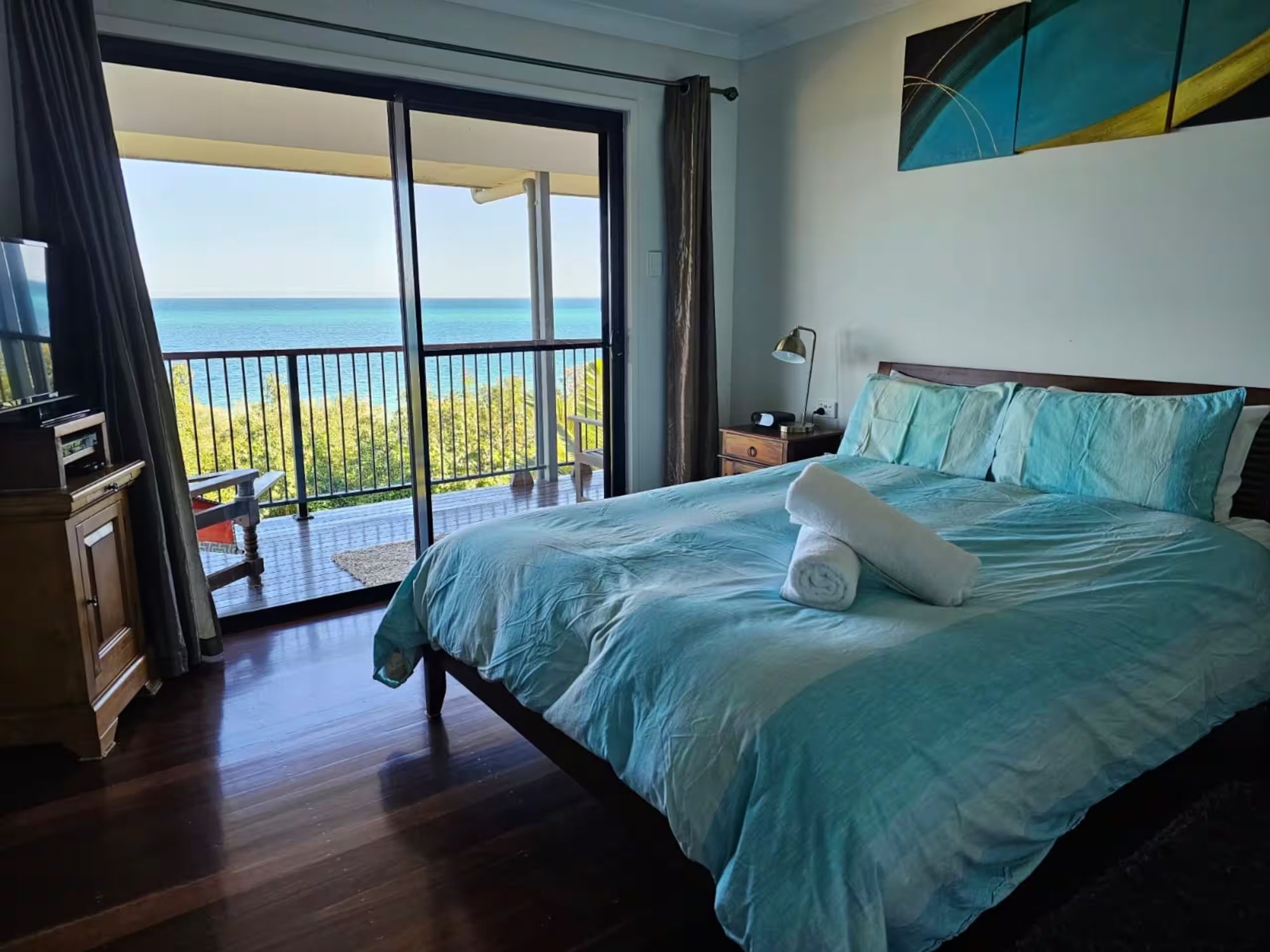 A hotel room with a large bed, a nightstand, a lamp, and a view of the ocean through a sliding glass door.
