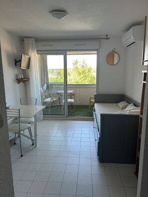 Habitación pequeña con cama, una mesita y una silla. Se ve un ventanal con vistas a zonas verdes y un edificio.