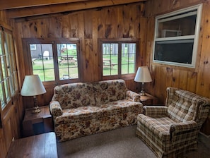 Ein Zimmer mit Holzwänden, einem blumigen Sofa, einem karierten Sessel, zwei Lampen und Blick auf einen Hinterhof mit einem Picknicktisch.