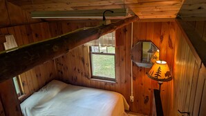Habitació petita i acollidora amb parets de fusta i sostre inclinat. Hi ha un llit individual amb matalàs blanc i una finestra petita amb vista a l'exterior. Una làmpada amb pantalla decorativa reposa sobre un prestatge de fusta.