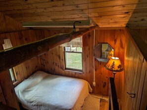 A small, cozy room with wooden walls and a slanted ceiling. There is a single bed with a white mattress and a small window with a view of the outdoors. A lamp with a decorative shade is on a wooden stand. - Honeymoon On The Water with private dock & Beach (Butternut)
