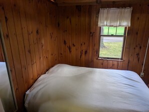 Ein Einzelbett in einem Zimmer mit Holzwänden und einem Fenster mit weißem Vorhang.