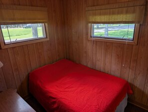 Ein Zimmer mit Holzwänden, einem Einzelbett mit roter Bettdecke und zwei Fenstern mit Jalousien.