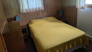 Ein Zimmer mit einem Bett, einem Nachttisch mit Lampe, einem Holzschrank und einem Fenster mit Vorhängen.