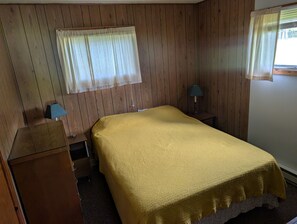 Ein Zimmer mit einem Bett, einem Nachttisch mit Lampe, einem Holzschrank und einem Fenster mit Vorhängen.