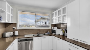A modern kitchen with white cabinets, a stainless steel dishwasher, a built-in microwave, and a window with blinds. - Bermagui Beach Apartment - Corunna Street 1 U2 (Bermagui)
