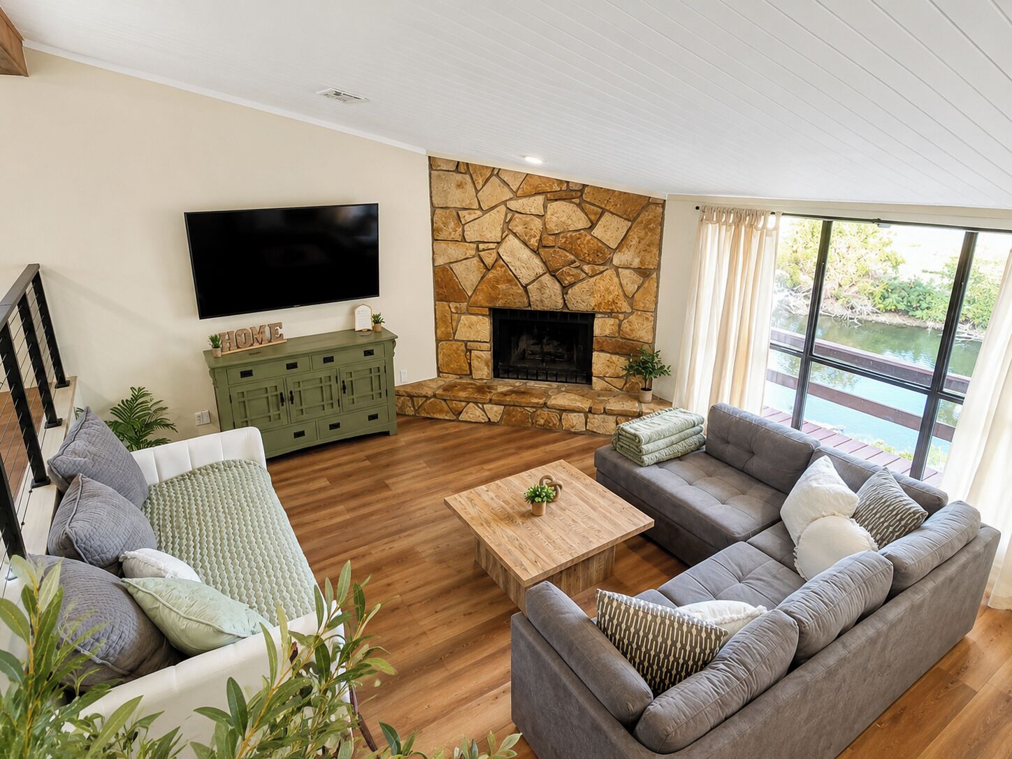 A modern living room with a stone fireplace, a flat-screen TV, a green cabinet, and a large window overlooking a body of water.