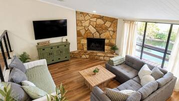 A modern living room with a stone fireplace, a flat-screen TV, a green cabinet, and a large window overlooking a body of water.