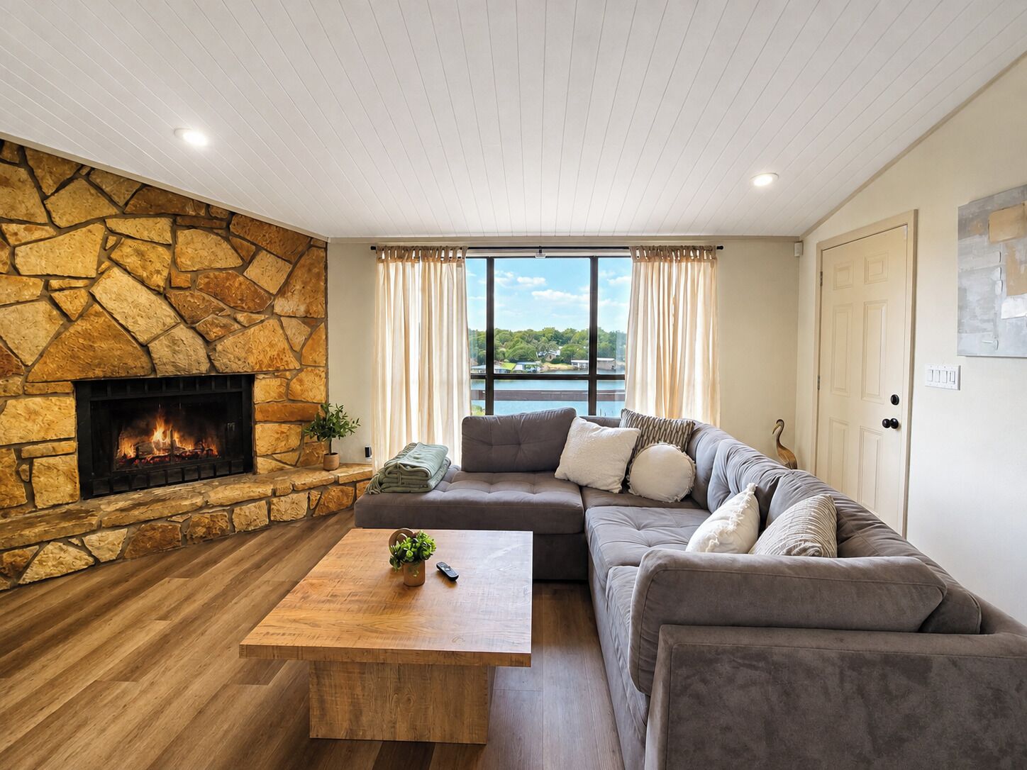 A living room with a stone fireplace, a large window with curtains, a gray sofa, and a wooden coffee table.