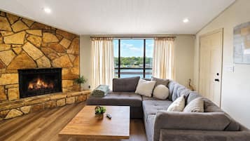 A living room with a stone fireplace, a large window with curtains, a gray sofa, and a wooden coffee table.