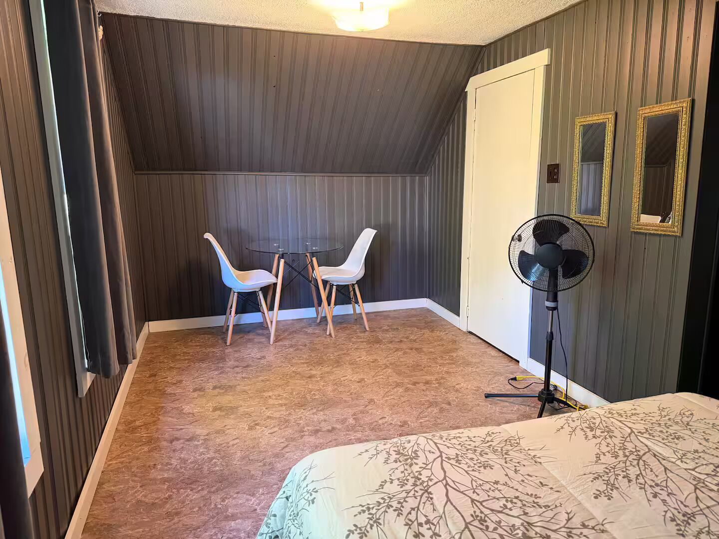 Ett rum med en säng, ett litet bord med två stolar, en golvfläkt och en dörr.