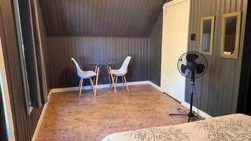 Ett rum med en säng, ett litet bord med två stolar, en golvfläkt och en dörr.