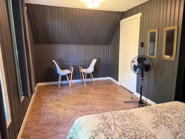 A room with a bed, a small table with two chairs, a standing fan, and a door.