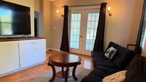 A living room with a dark sofa, a round wooden coffee table, a flat-screen TV on a wooden cabinet, and a sliding glass door with curtains.