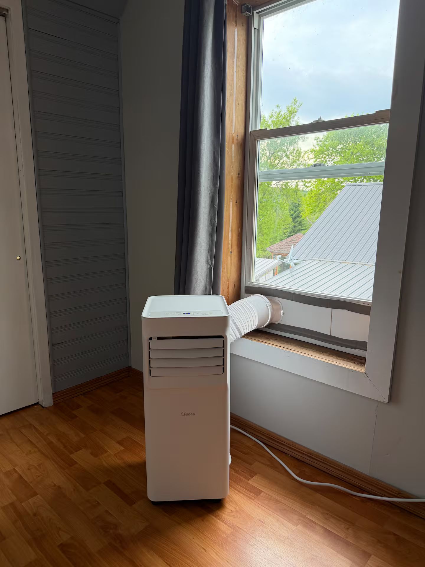 A room with a window, a white air conditioner, and a wooden floor.
