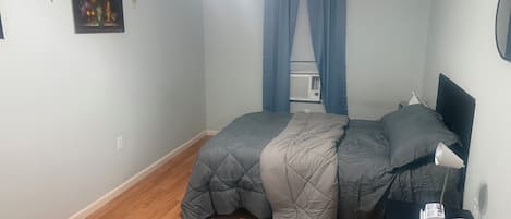 Un dormitorio con cama, mesita de noche, lámpara, ventana con cortinas y aire acondicionado instalado en la pared.