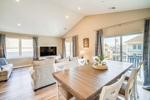 A spacious living area with a wooden dining table, white chairs, a flat-screen TV, and large windows offering a view of the city. - Fantastic 4-bedroom condo - Walk to the Beach! (Wildwood)