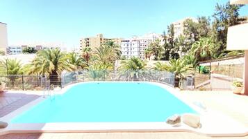 A swimming pool with a surrounding fence, palm trees, and buildings in the background.