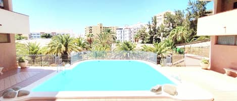 A swimming pool with a surrounding fence, palm trees, and buildings in the background.