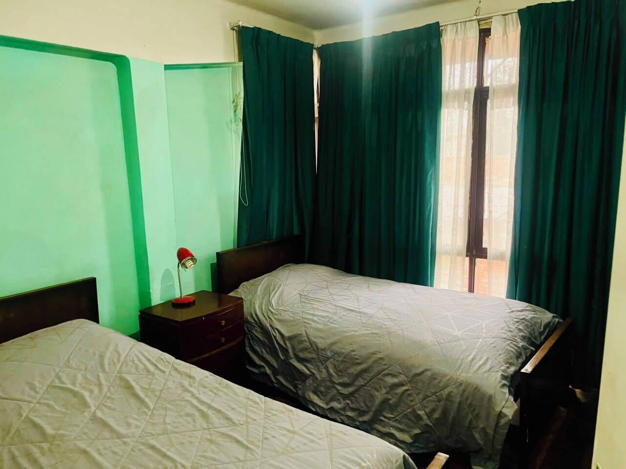 A room with two beds, a nightstand with a lamp, and a window with curtains.