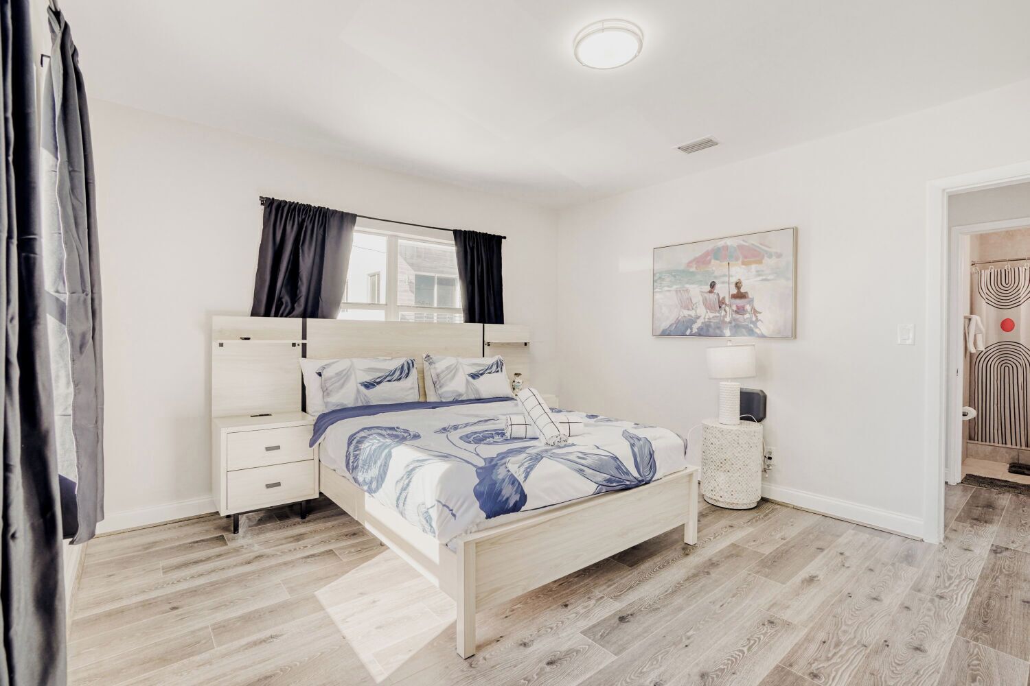 A bedroom with a bed, bedside tables, a nightstand, a window with curtains, a wall-mounted picture, and a door leading to the bathroom.