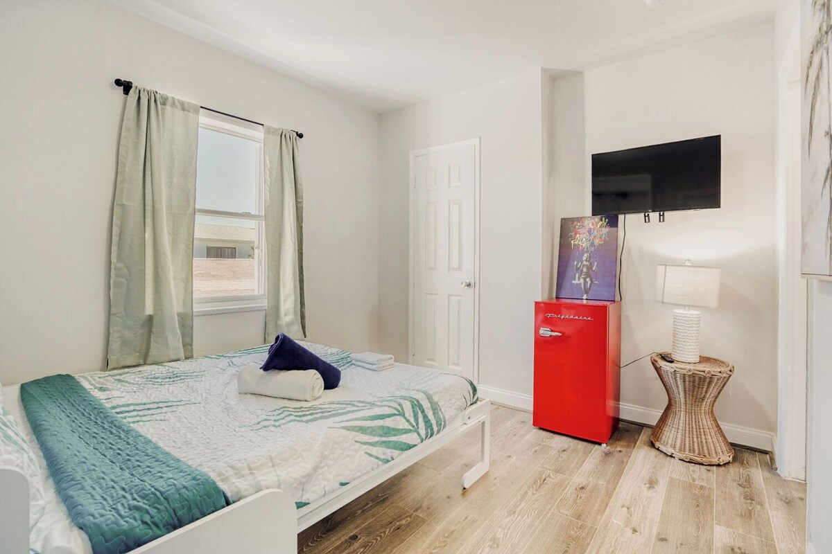 A hotel room with a bed, a television mounted on the wall, a small red refrigerator, and a wicker side table with a lamp.