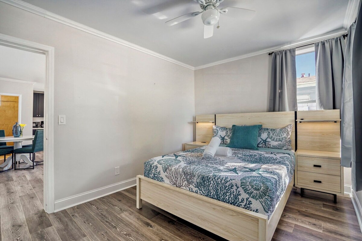 A bedroom with a bed, a nightstand, a ceiling fan, and a view of the outside.