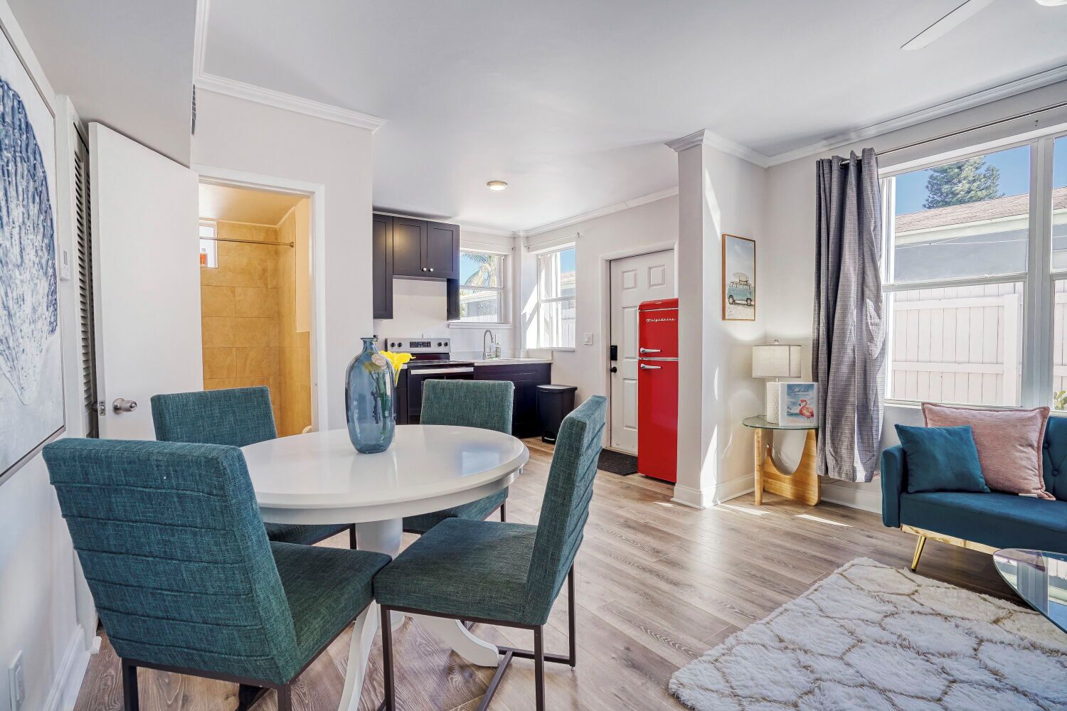 A modern living room with a round dining table, teal chairs, a red refrigerator, and a large window with curtains.