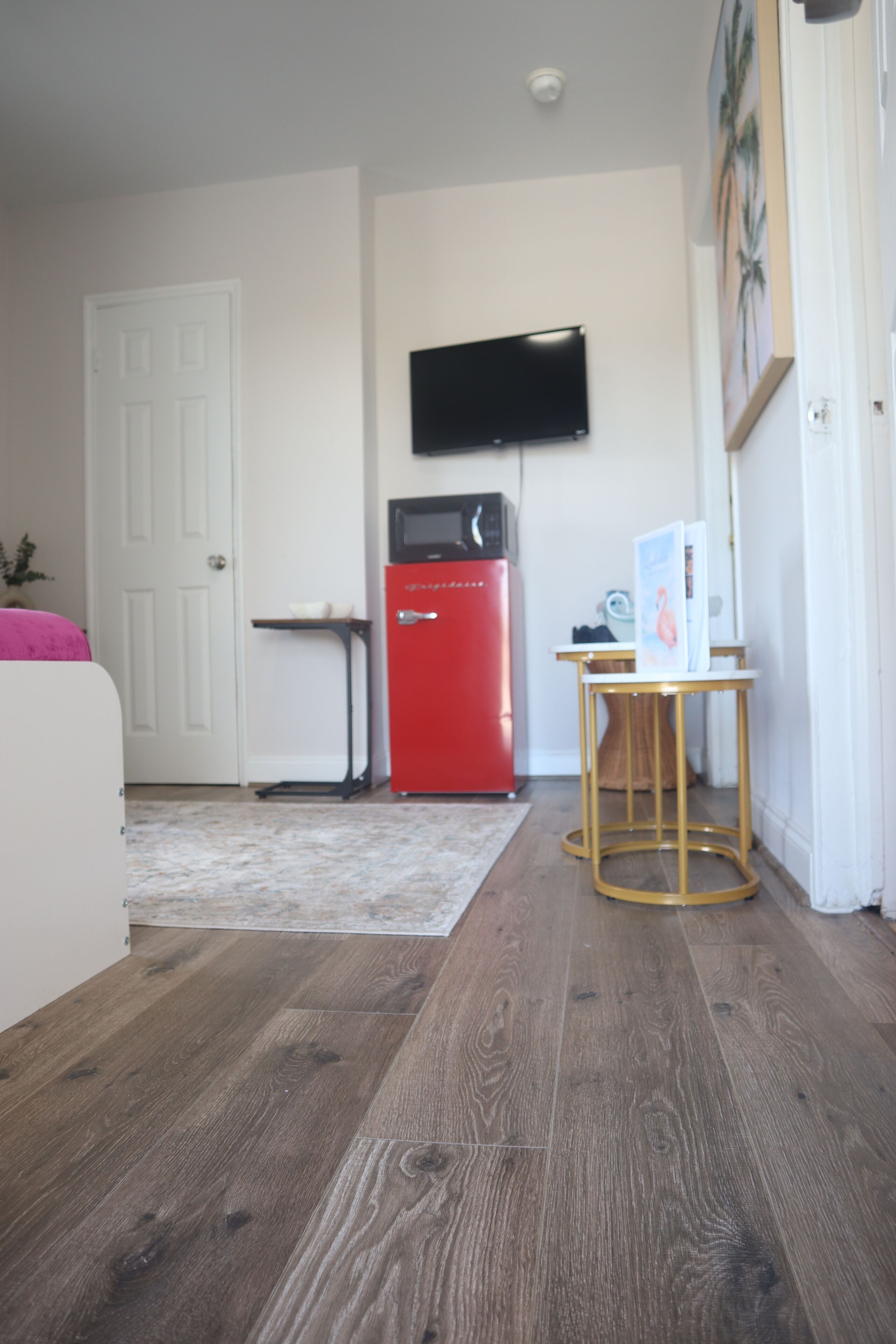 Una habitación con piso de madera, una puerta blanca, un electrodoméstico rojo, un televisor de pantalla plana montado en la pared y una mesita con una revista.
