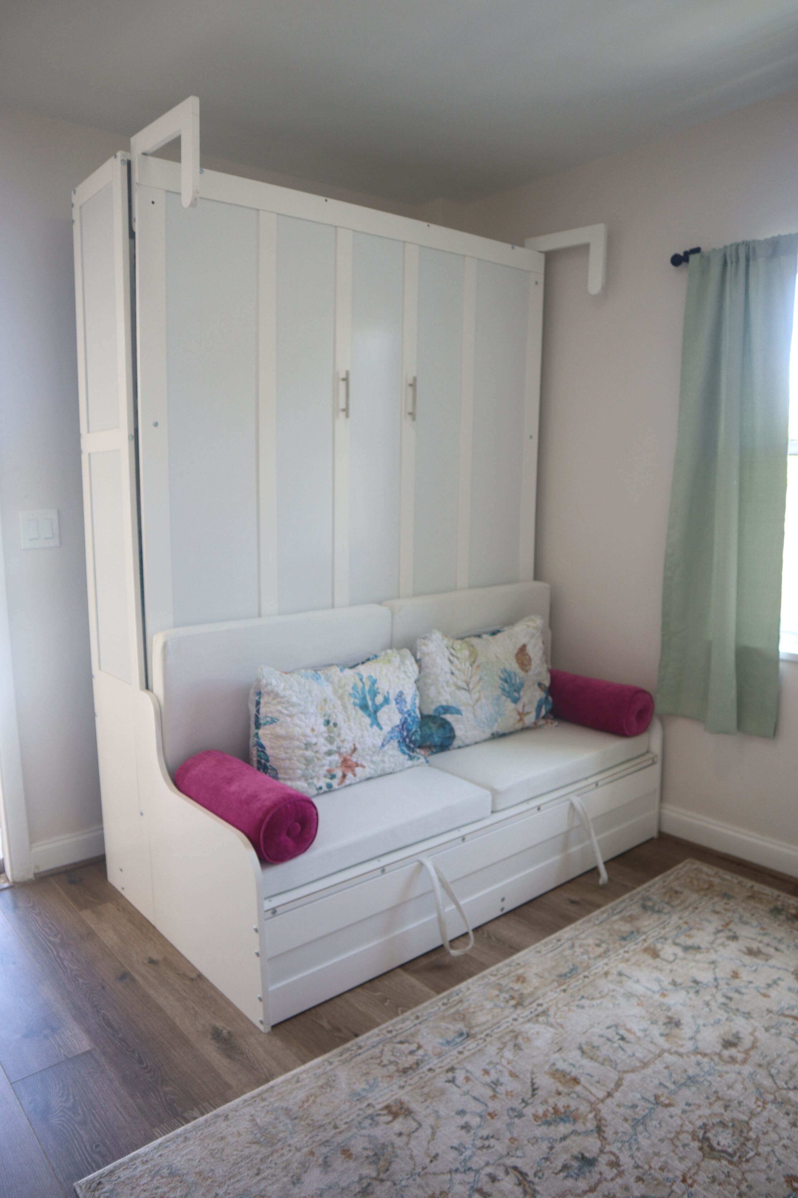 Un sofá blanco con un cojín floral y una toalla rosa, frente a una pared con cortina verde claro.