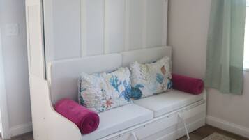 Un sofá blanco con un cojín floral y una toalla rosa, frente a una pared con cortina verde claro.