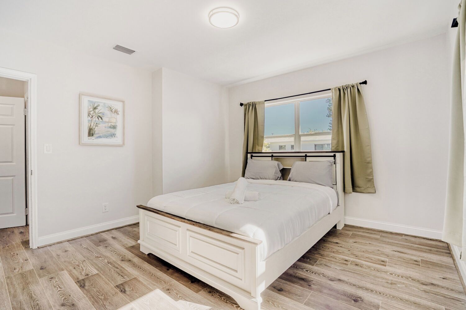 Un dormitorio con cama, ventana con cortinas, puerta y un cuadro colgado en la pared.