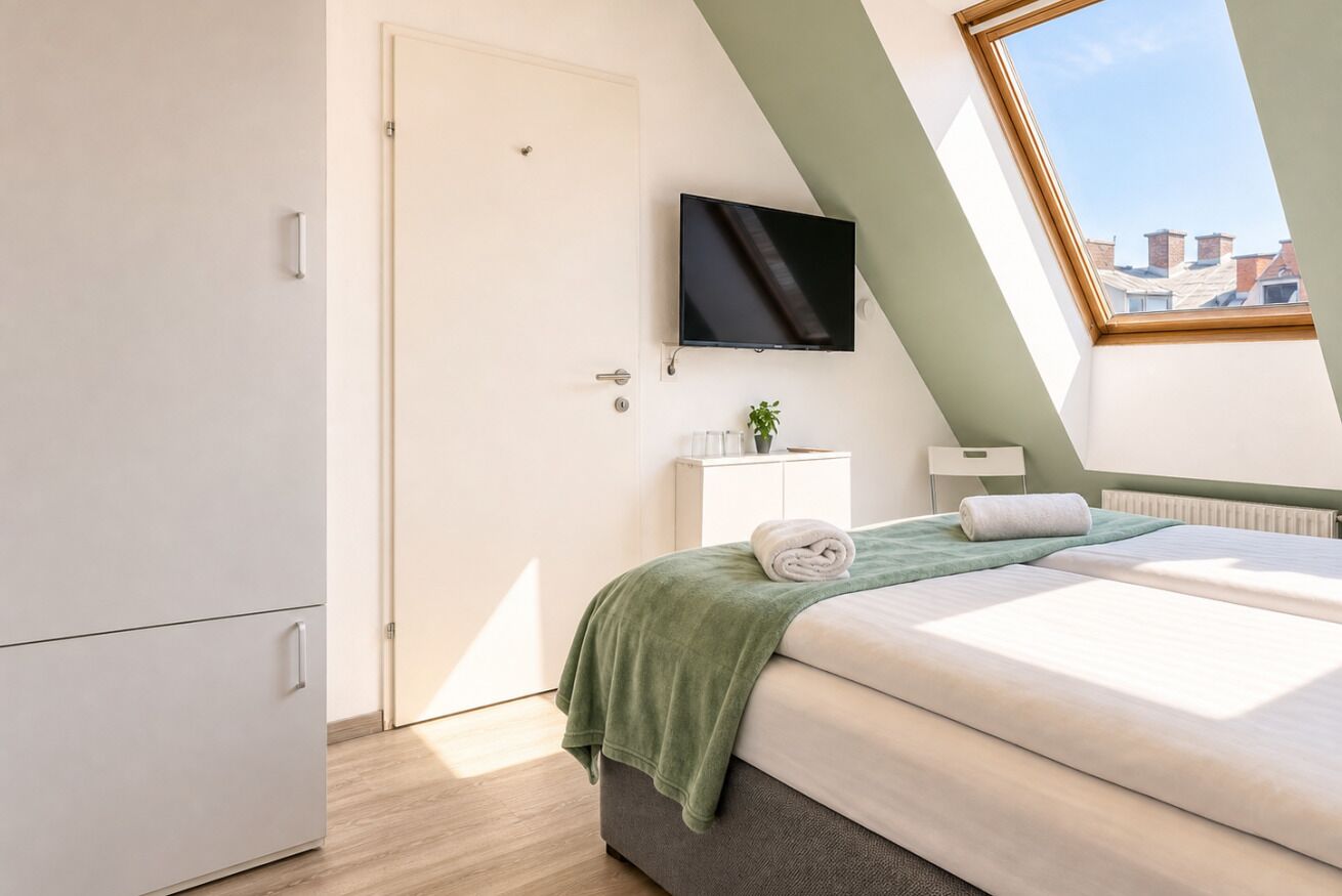 Une chambre à coucher avec un lit, un téléviseur fixé au mur, un radiateur et une fenêtre donnant sur des immeubles.