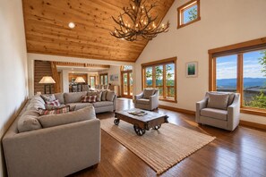 A spacious living room with a wooden ceiling, a fireplace, and large windows offering a view of the outdoors. - Kumbaya House: Steps from Cupp Run | Hot Tub, Deck & Epic Sunset Views (Snowshoe)