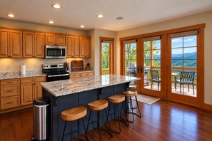 A kitchen with wooden cabinets, a granite countertop, a stainless steel refrigerator, and a large island with bar stools. - Kumbaya House: Steps from Cupp Run | Hot Tub, Deck & Epic Sunset Views (Snowshoe)