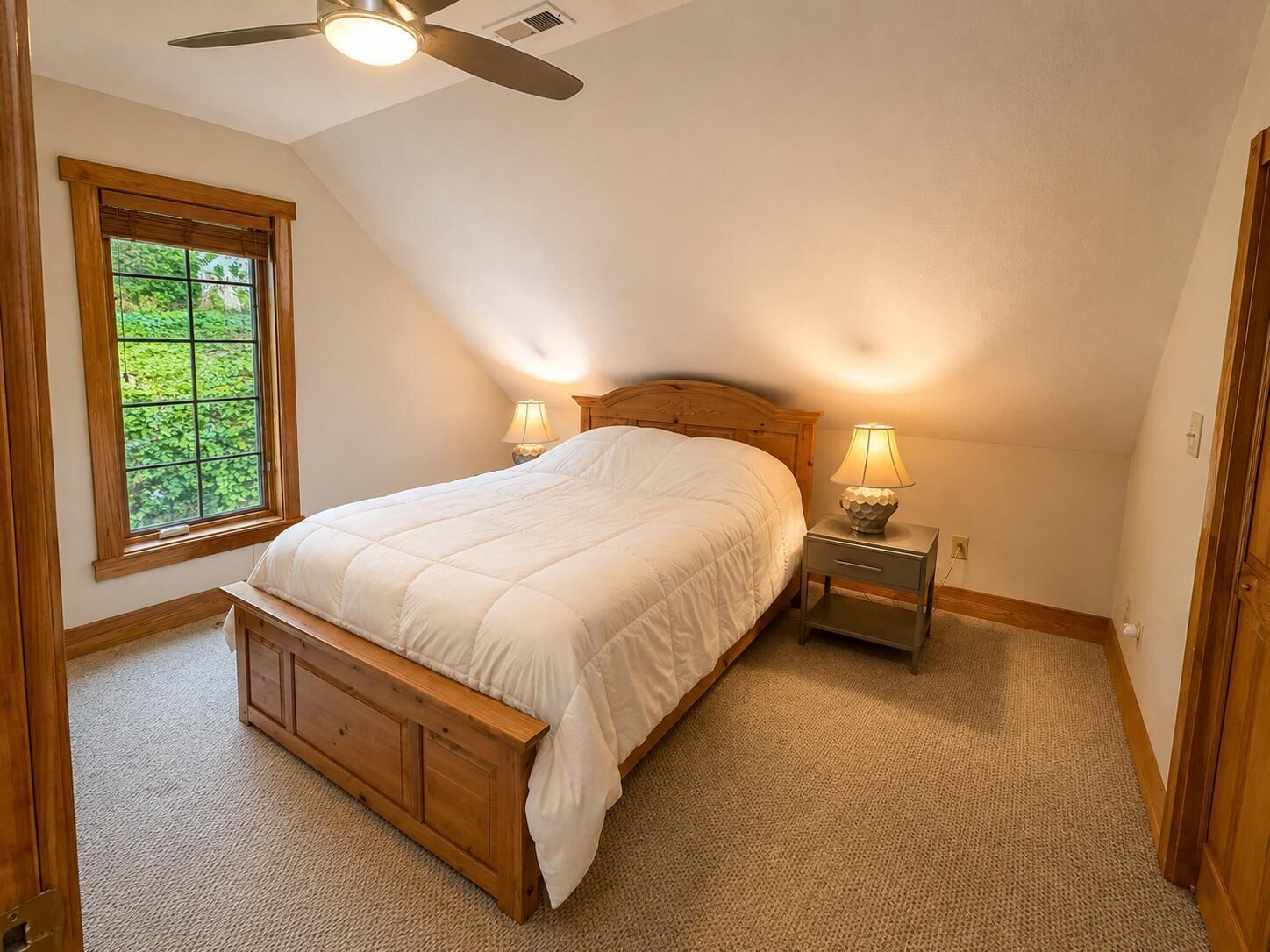 A bedroom with a bed, two bedside tables with lamps, a ceiling fan, and a window with a view of greenery.