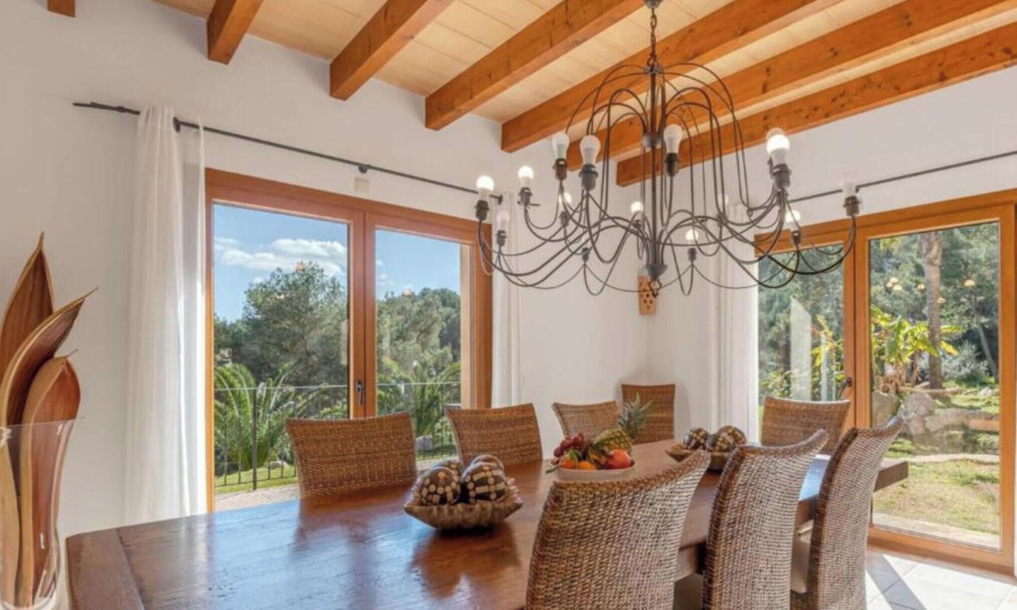 A dining area with a wooden table, wicker chairs, a chandelier, and large windows with a view of trees.