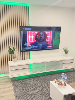 Televisão exibindo um programa com esquema de cores vermelho e preto, um móvel de entretenimento branco, uma área iluminada em verde e uma pequena planta sobre o móvel.